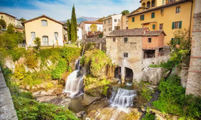 Cascata e mulino in pietra, tra i borghi della Toscana. Case gialle e verdi colline sullo sfondo.
