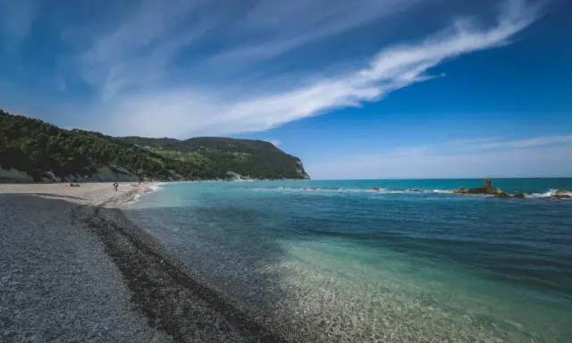 Spiaggia di ciottoli con mare cristallino e una collina verdeggiante, tipica del entroterra marchigiano.
