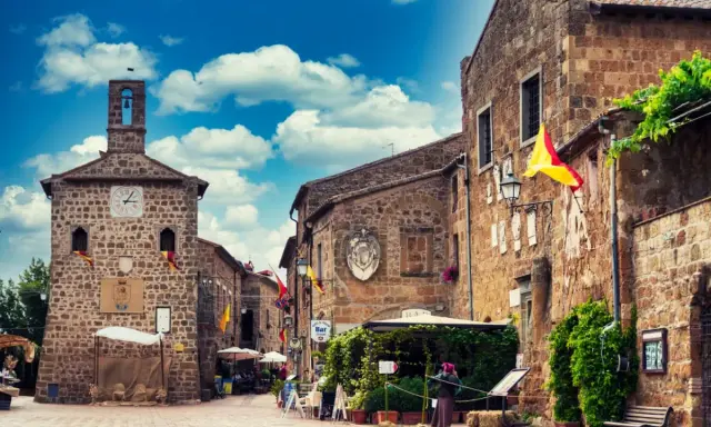Un vicolo pittoresco a Sovana, Italia, con edifici in pietra, bandiere colorate e un cielo azzurro pieno di nuvole.