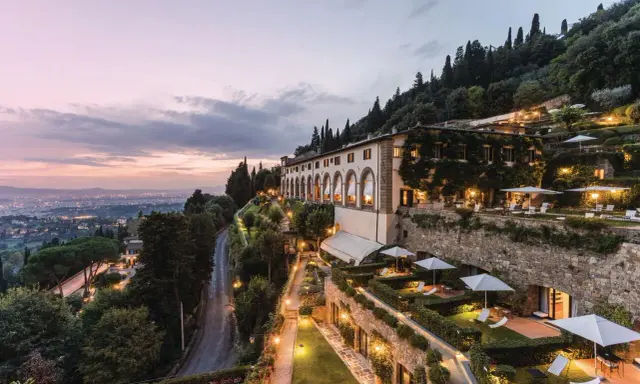 Un lussuoso hotel si affaccia sulle **colline di Firenze** al tramonto, con giardini illuminati e la città in lontananza.