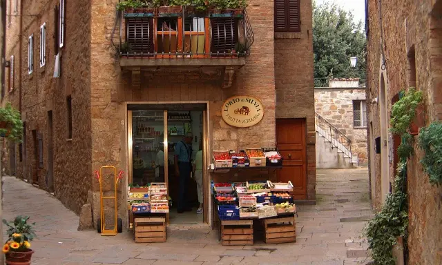 Un piccolo negozio di frutta e verdura in un vicolo di **paesi toscani**. Frutta fresca in cassette di legno, un balcone fiorito e l'atmosfera autentica.