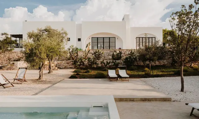 Piscina e lettini in una delle masserie famose puglia, con ulivi e cactus sotto un cielo azzurro con nuvole bianche.