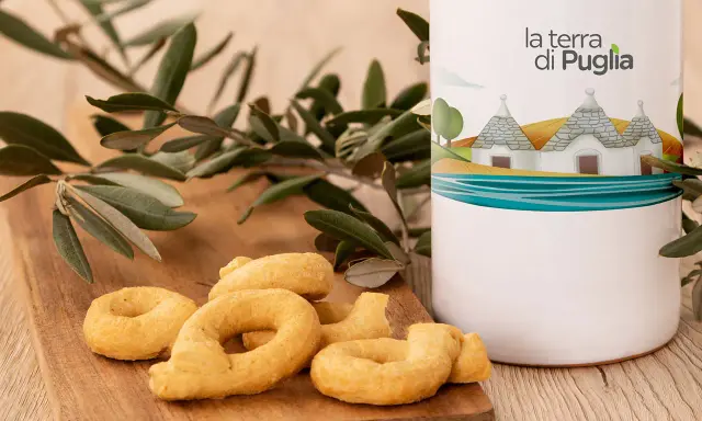 Taralli pugliesi su tagliere di legno, accanto a un contenitore con il logo "la terra di Puglia" e un ramo d'ulivo.