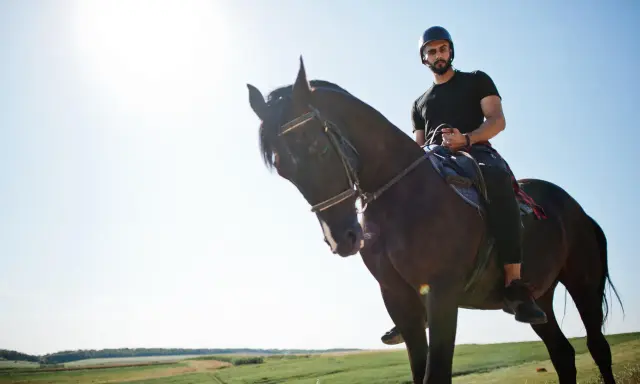 Uomo con casco e barba cavalca un cavallo scuro in un paesaggio verdeggiante. Un'esperienza di **Toscana a cavallo**.