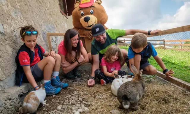 Bambini e adulti interagiscono con conigli in un recinto, parte di un'esperienza in fattorie didattiche. Un personaggio con costume da orso sorride.