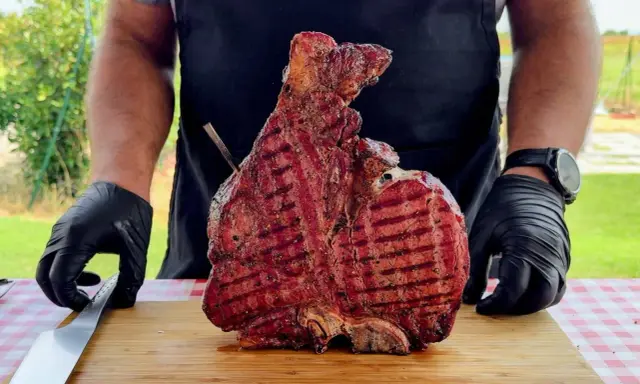 Un cuoco con guanti neri mostra una succulenta bistecca alla fiorentina appena grigliata, pronta per essere servita. Un esempio perfetto di come cucinare la bistecca alla fiorentina.