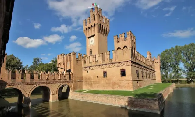 Un imponente castello in mattoni rossi, uno dei tanti castelli Emilia Romagna, con un ponte ad archi sull'acqua e torri merlate sotto un cielo azzurro.