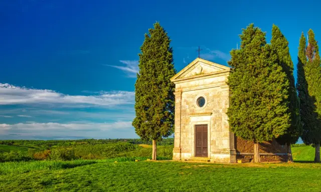 Cappella di San Quirico d'Orcia, circondata da cipressi, su un prato verde sotto un cielo azzurro.