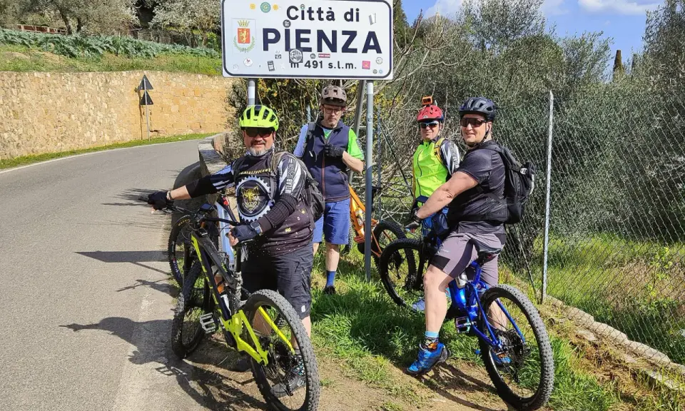Quattro ciclisti si fermano per una foto con il cartello "Città di Pienza" durante un'avventura in bici nel Val d'Orcia.