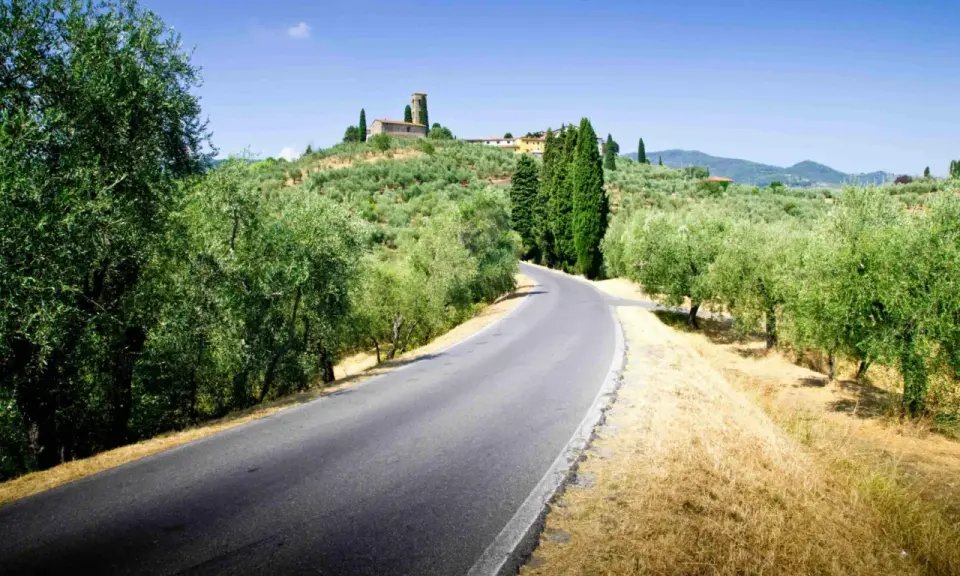 Strade del vino toscana: una curva su un asfalto grigio, circondata da ulivi e cipressi, con un borgo in collina.