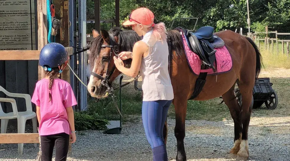 Bambina con casco blu e maglietta rosa osserva un cavallo marrone pronto per un giro a cavallo per bambini. Una ragazza con cappellino arancione prepara il cavallo.