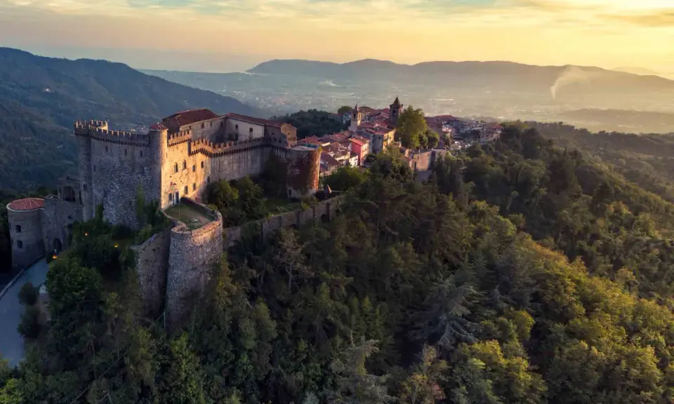 Antico castello medievale su una collina boscosa, uno dei tanti castelli da visitare in Toscana, con un villaggio arroccato e vista sulla valle al tramonto.