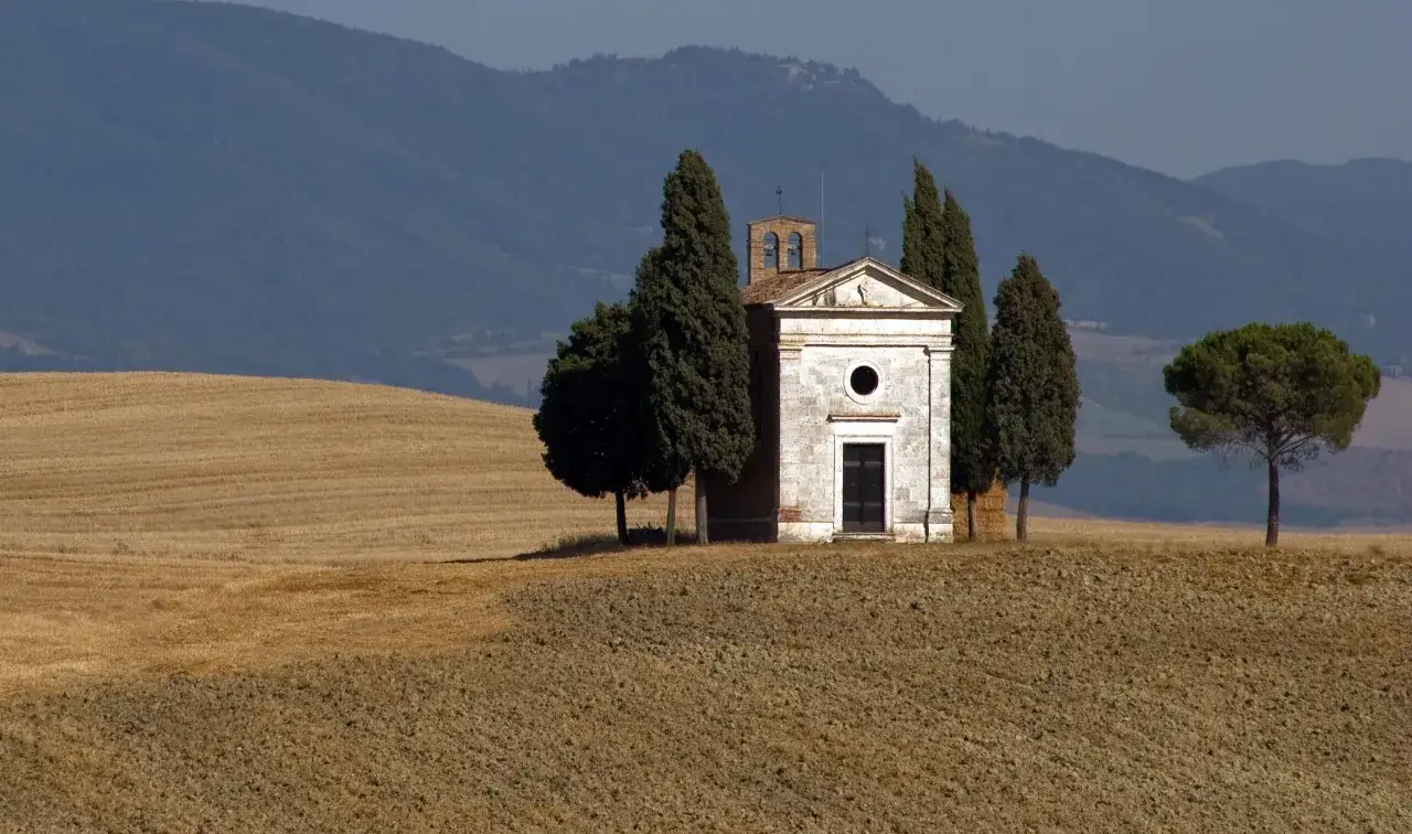 La chiesa di Vitaleta, circondata da cipressi e campi dorati, si erge solitaria sotto un cielo azzurro.