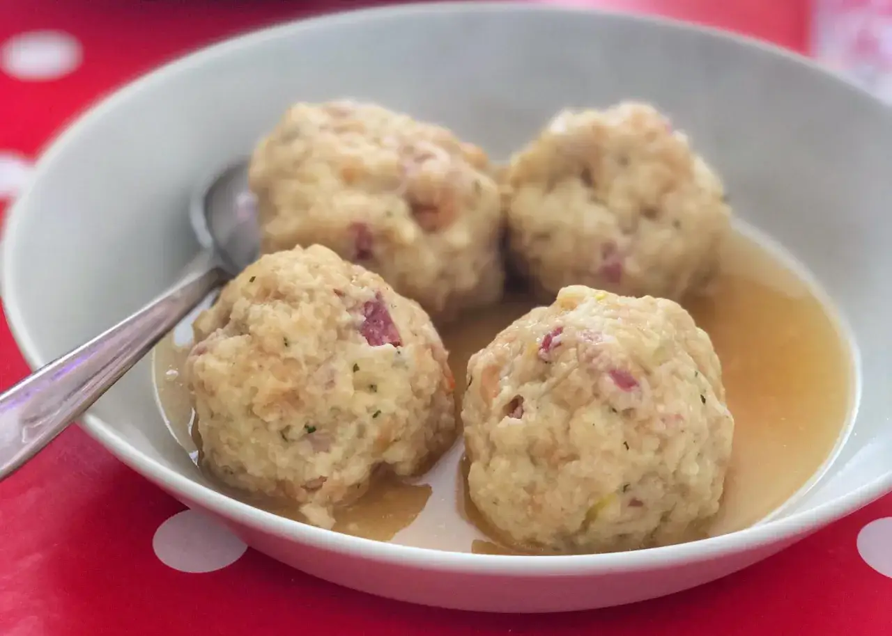 Canederli in brodo, un classico dei cibi tipici del Trentino Alto Adige, serviti in una ciotola bianca su un tavolo rosso a pois bianchi.