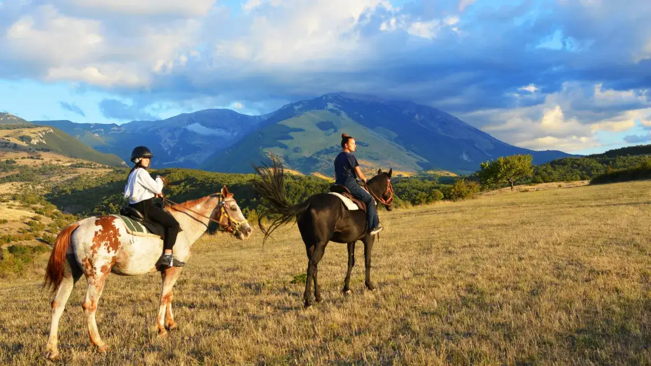 Due persone godono di passeggiate a cavallo in Abruzzo, con montagne maestose sullo sfondo e un cielo drammatico.