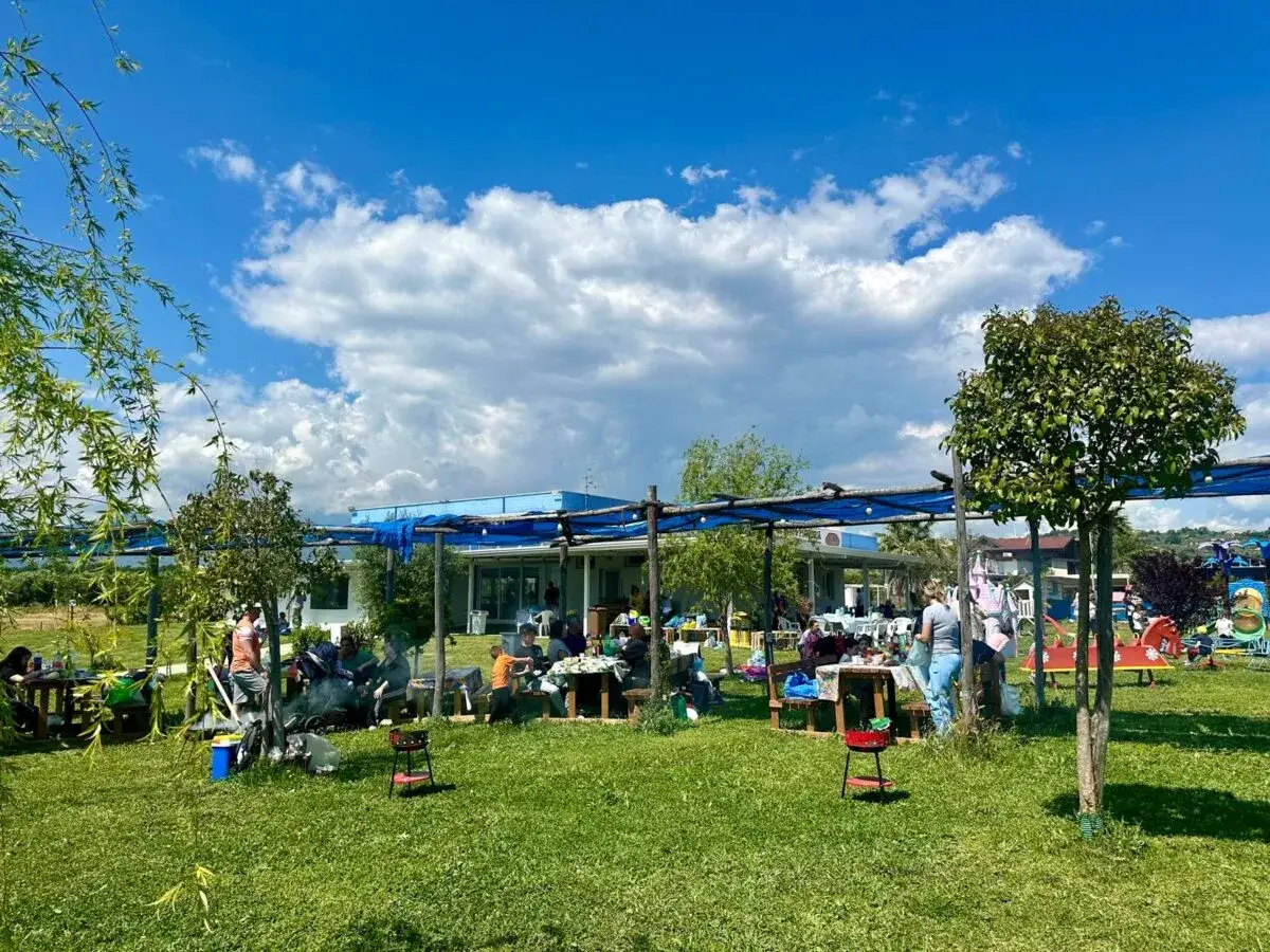Famiglie riunite in un prato verde sotto un cielo azzurro con nuvole bianche. Un luogo ideale tra i migliori agriturismi per bambini in Campania, con aree picnic e giochi.