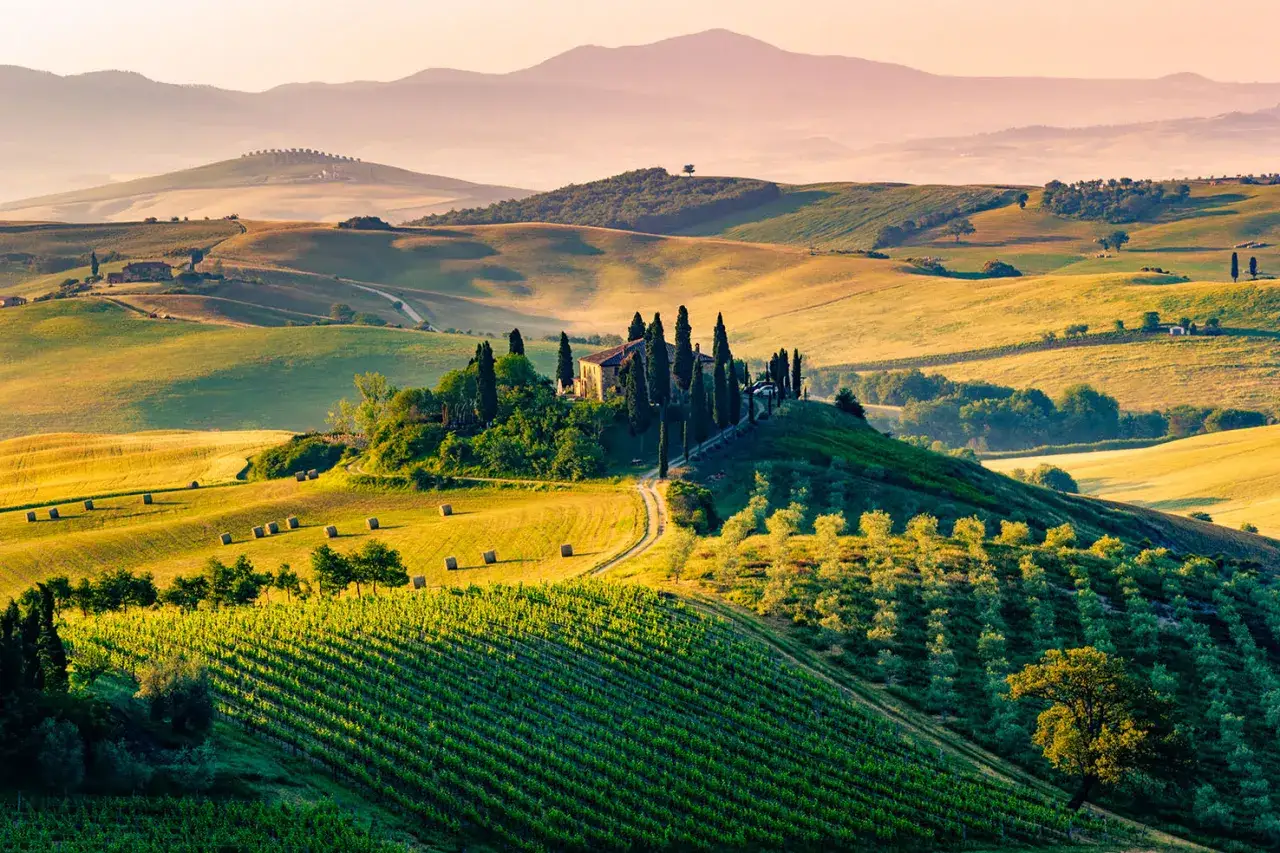 Dolci colline del Val d'Orcia in primavera, con un casale in cima a una collina circondato da cipressi e vigneti rigogliosi.