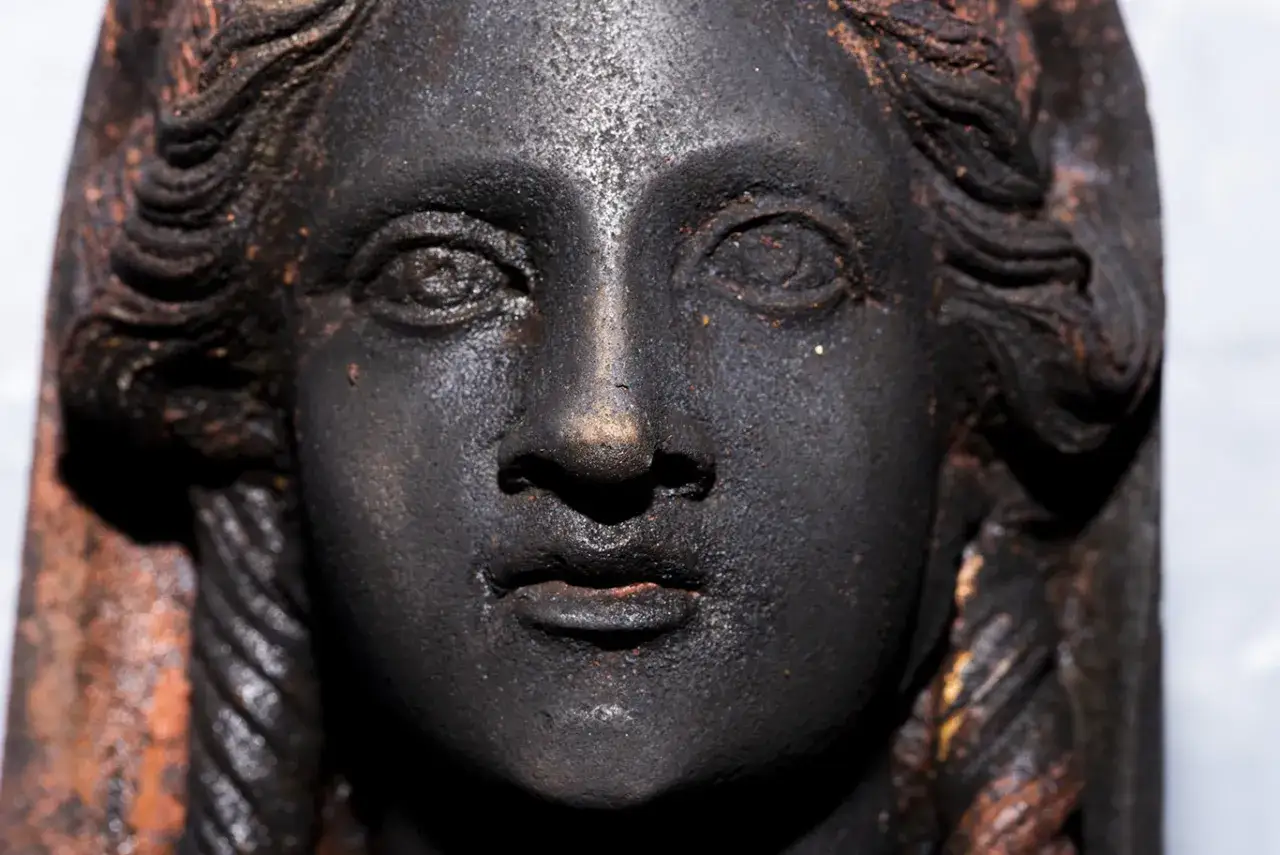Volto di bronzo scuro, un reperto dei bronzi di San Casciano dei Bagni, con capelli a onde e trecce.