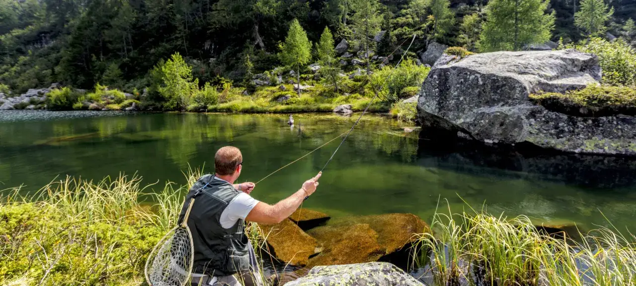 Uomo pratica pesca a mosca in un lago alpino del Trentino Alto Adige, circondato da rocce e fitti boschi.