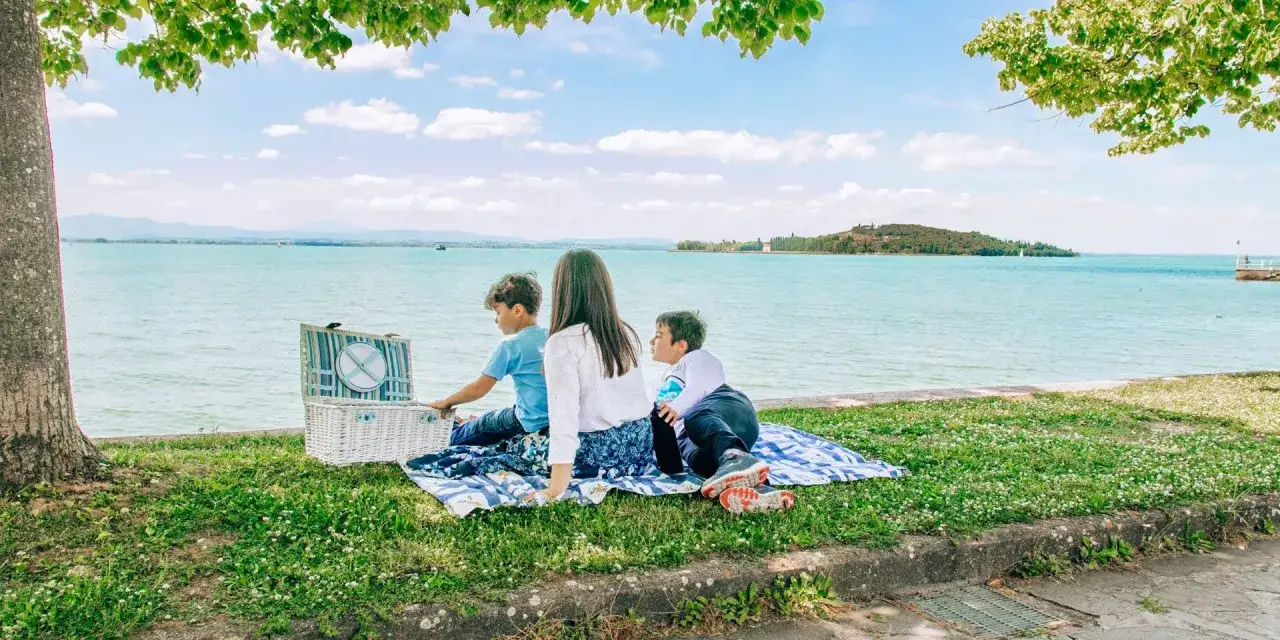 Picnic con bambini al lago Trasimeno. Una famiglia si gode la giornata con un cestino da picnic e una coperta sull'erba, con il lago sullo sfondo.