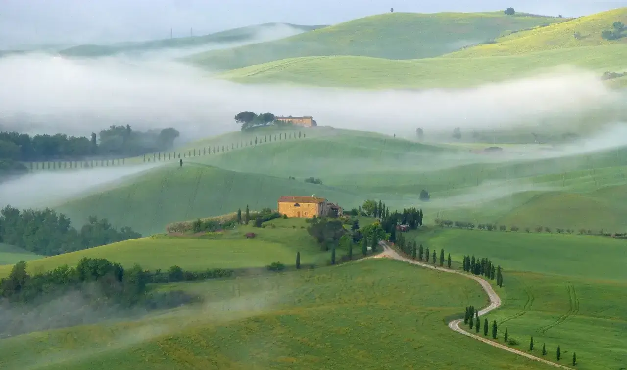 Dolci colline verdi avvolte dalla nebbia mattutina nel Val di Norcia, con casali in pietra e filari di cipressi.