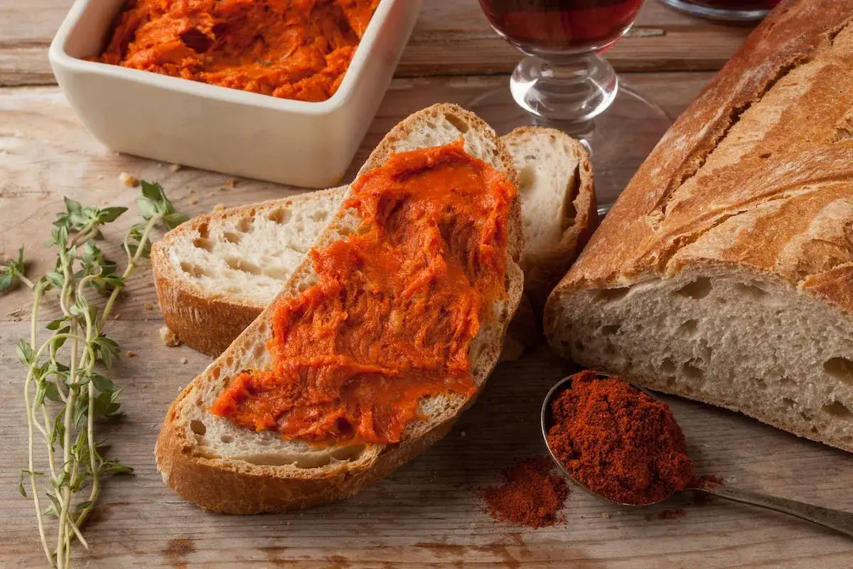 Fetta di pane con 'nduja, un assaggio di vero mangiare calabrese. Accanto, pane fresco, erbe aromatiche e un bicchiere di vino rosso.