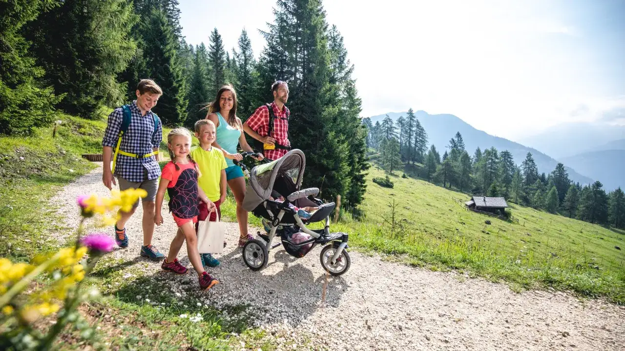 Famiglia in escursione in montagna per bambini estate, con passeggino e bambini felici su sentiero tra alberi e prati verdi.
