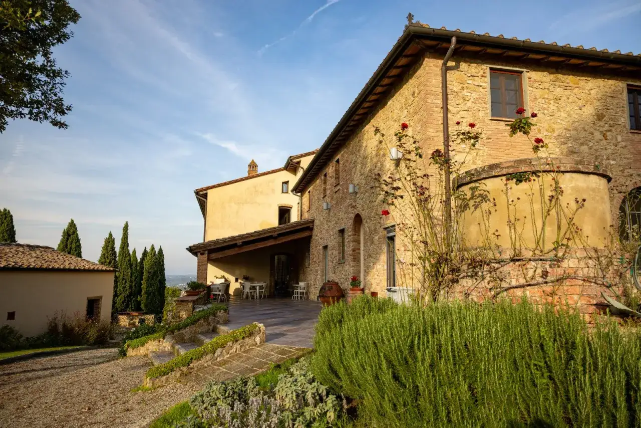 Un casale in pietra dorata, con rose rosse che si aggrappano al muro, incornicia un paesaggio collinare. La Toscana in inverno mostra la sua bellezza austera sotto un cielo azzurro.
