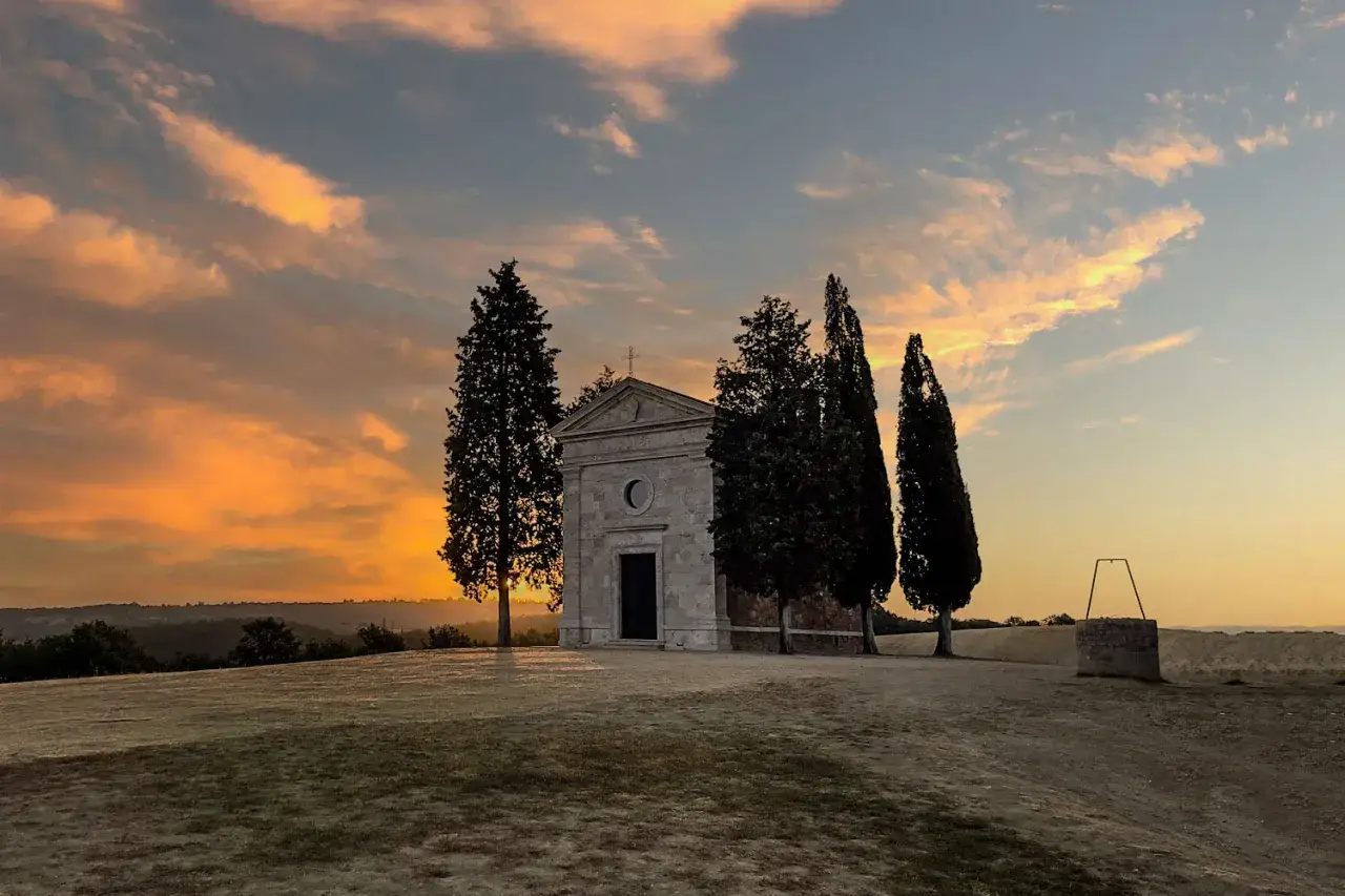 La cappella della Madonna di Vitaleta, circondata da cipressi, si staglia contro un cielo infuocato al tramonto.