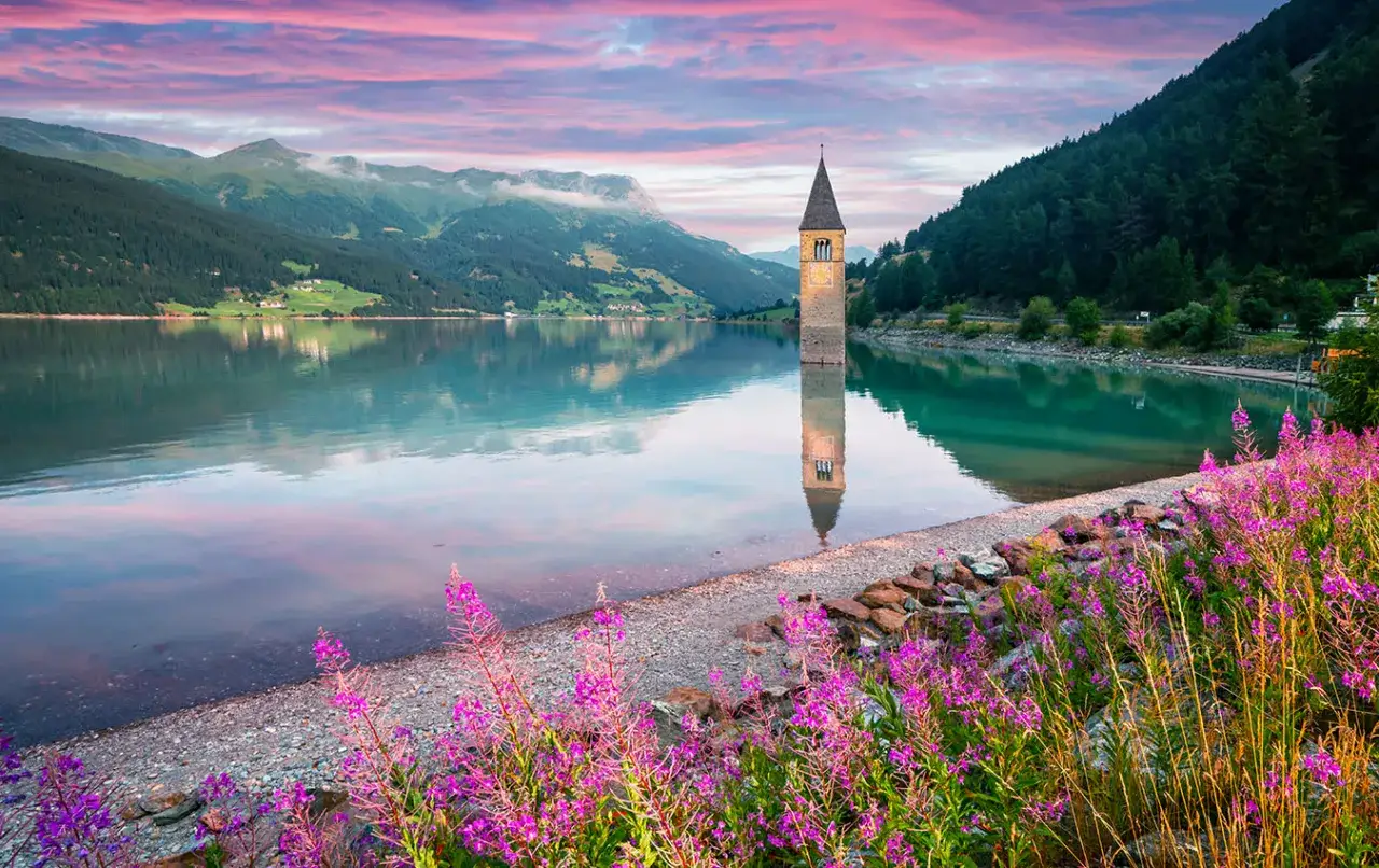 Torre sommersa in un lago alpino, con montagne verdi e cielo rosa al tramonto. Un angolo di montagna Friuli Venezia Giulia incantevole.