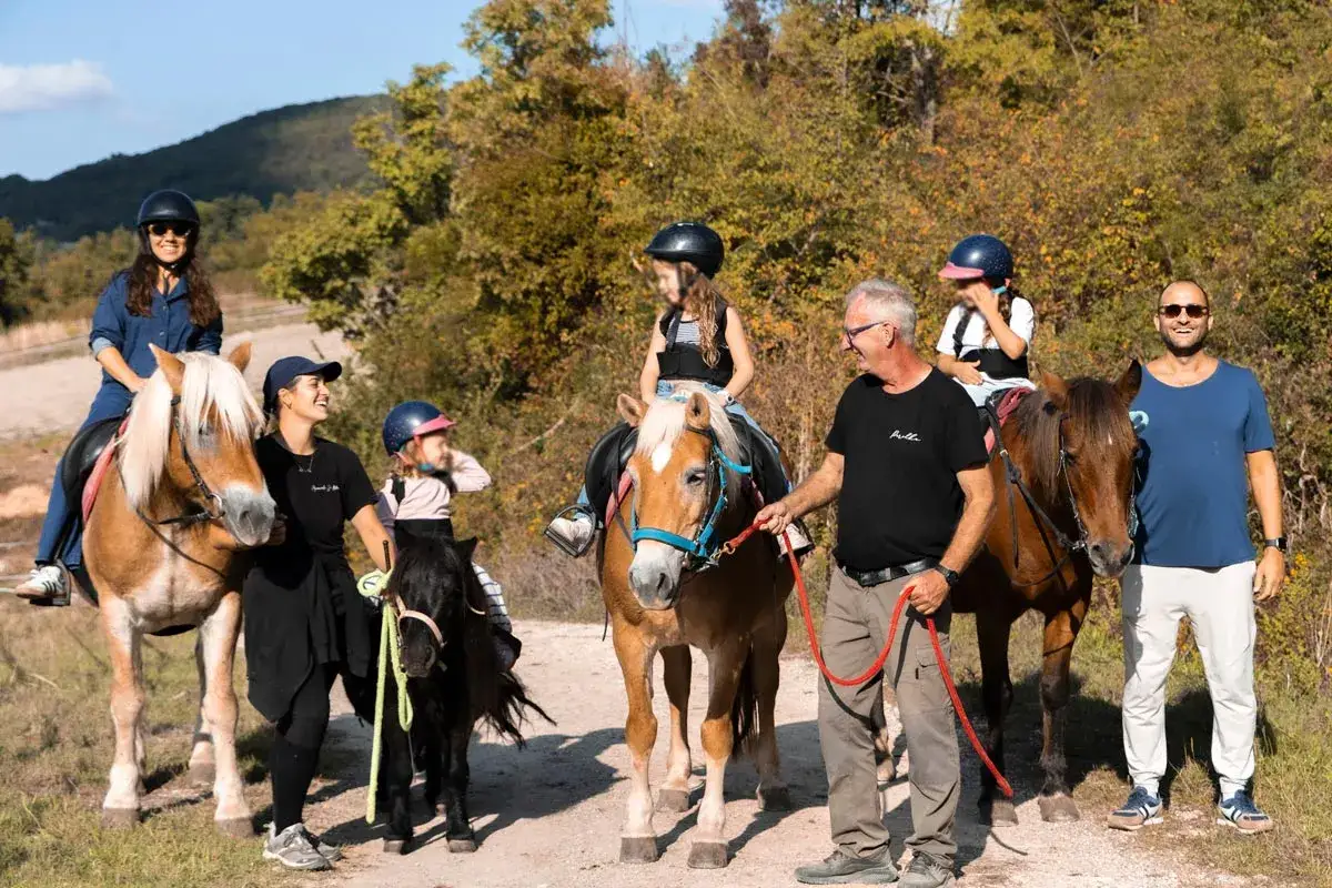 Famiglia in gita in un hotel con maneggio, bambini e adulti a cavallo e pony, in una giornata di sole tra alberi autunnali.