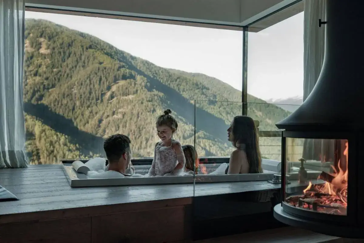 Una famiglia si gode un bagno caldo in una vasca con vista sulle montagne. Un'esperienza di spa per famiglie indimenticabile, con un camino acceso per un'atmosfera accogliente.
