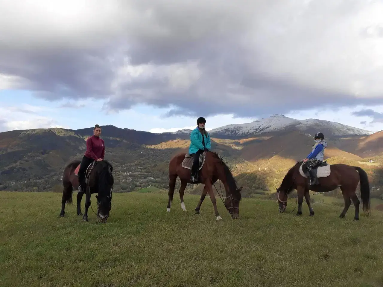 Tre persone a cavallo godono di passeggiate a cavallo in Emilia Romagna, con montagne innevate sullo sfondo.