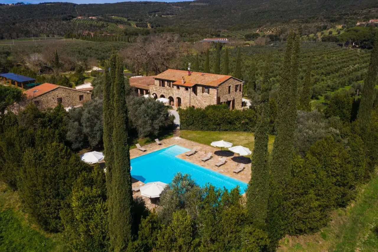 La campagna toscana lungo il mare, con un casale in pietra, piscina azzurra e cipressi svettanti tra uliveti.