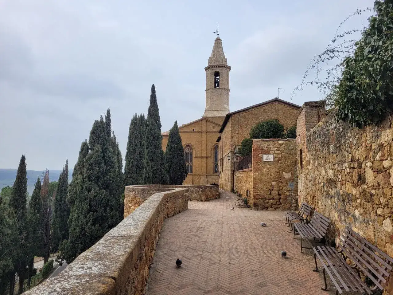 Un vicolo di Pienza, con la sua storia rinascimentale, fiancheggiato da cipressi e antiche mura in pietra. Panchine invitano a contemplare il paesaggio.