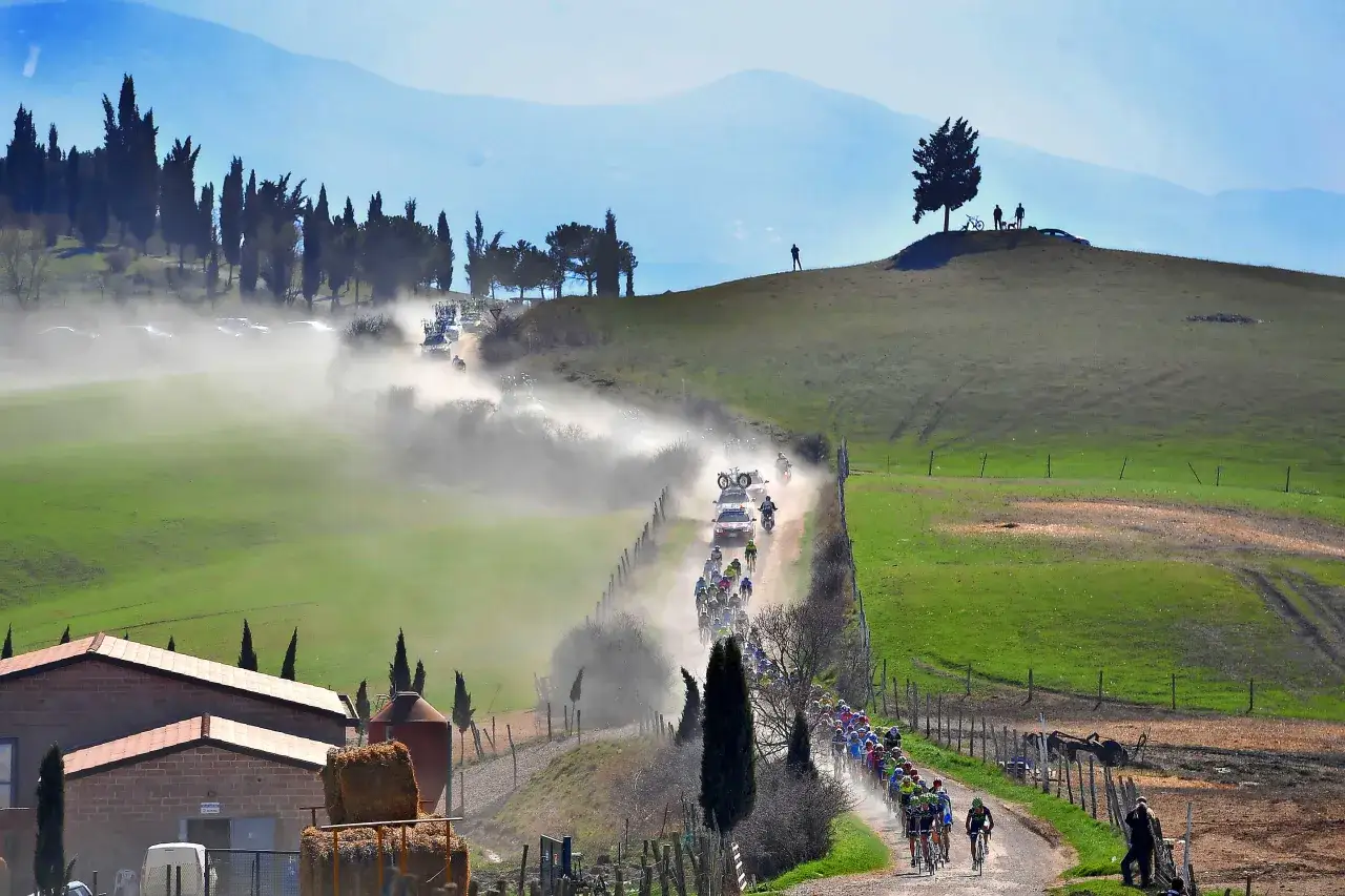 Un gruppo di ciclisti pedala su una strada sterrata in Toscana, sollevando polvere tra colline verdi e cipressi.