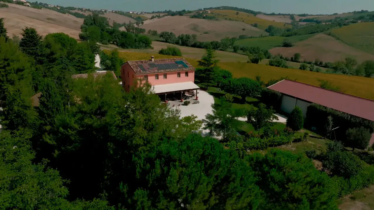 Agriturismi colline marchigiane: un casale rosa con pannelli solari, circondato da alberi e campi di girasoli, immerso nel verde.