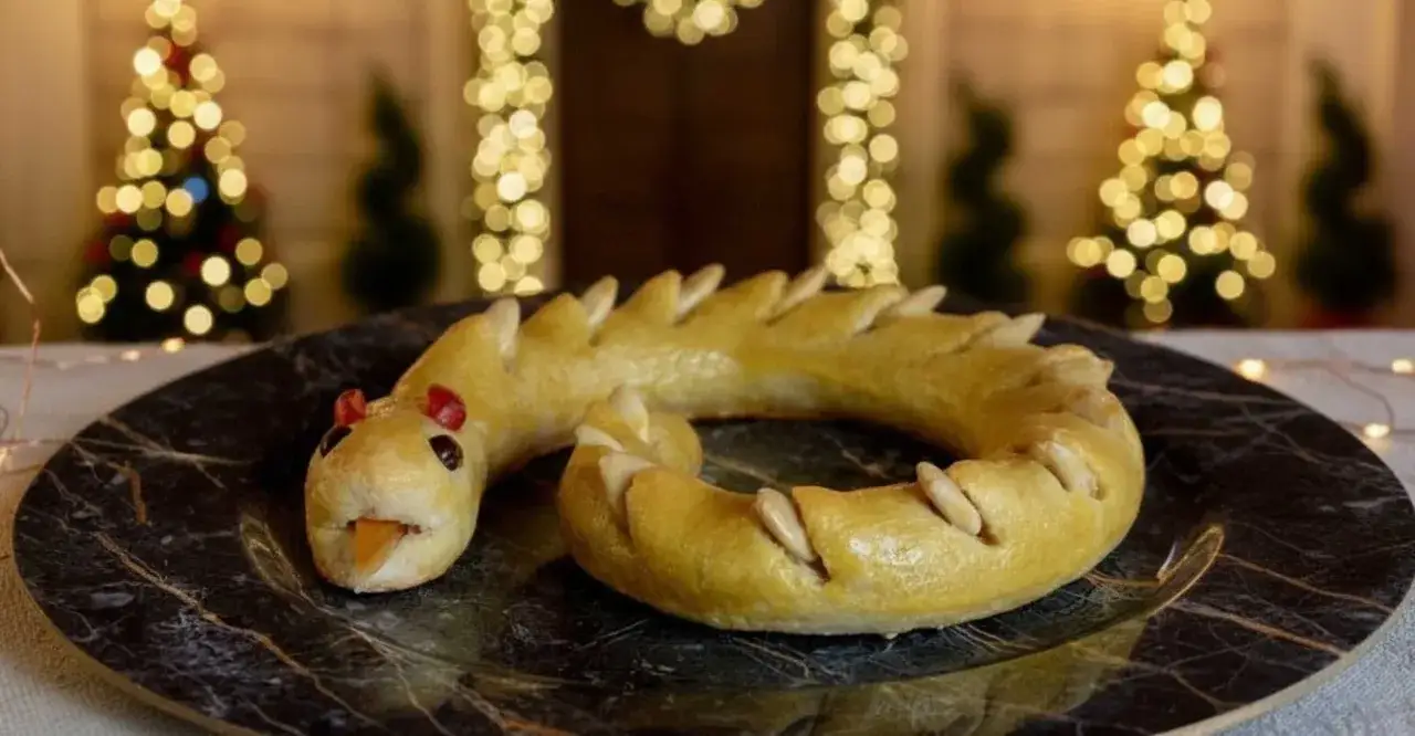 Dolci tipici Perugia: un serpente di pasta frolla dorata, decorato con mandorle e bacche rosse, su un piatto di marmo, con alberi di Natale sfocati sullo sfondo.