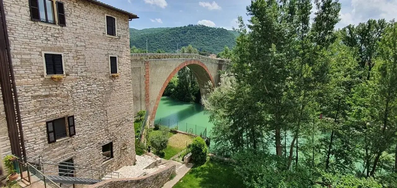 Un antico ponte ad arco domina un fiume verde smeraldo, circondato da lussureggianti colline. Perfetto per le tue prossime escursioni nelle Marche.