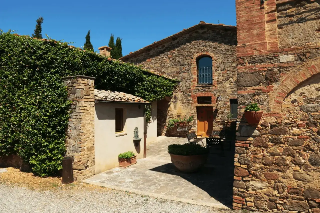 Cortile di antichi paesi toscani, con muri in pietra e mattoni, edera rampicante e un cielo azzurro intenso.
