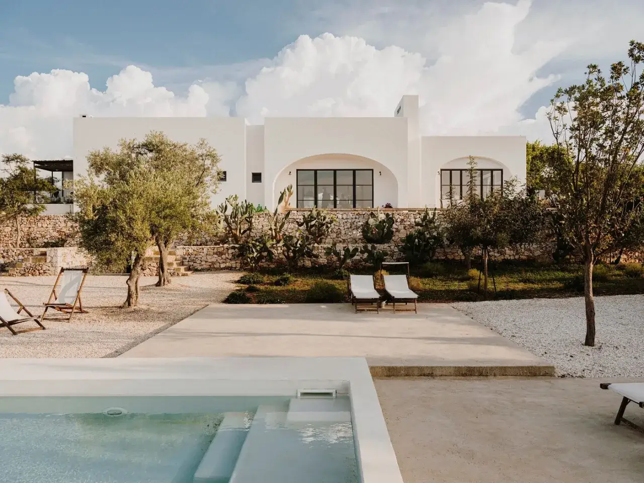Piscina e lettini in una delle masserie famose puglia, con ulivi e cactus sotto un cielo azzurro con nuvole bianche.