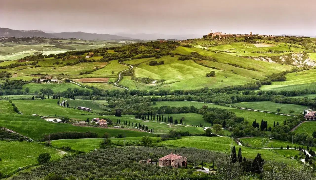 Dolci colline verdi con un borgo toscano da visitare in lontananza. Campi coltivati, uliveti e cipressi disegnano un paesaggio incantevole.