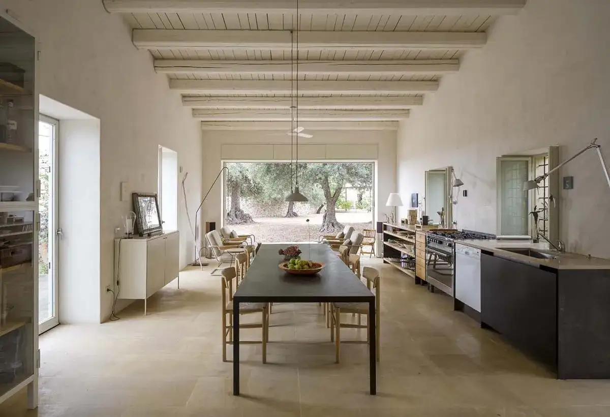 Interno di una casa con travi a vista e un grande tavolo da pranzo, che si affaccia su un giardino con ulivi. Un'atmosfera da bagli in Sicilia.