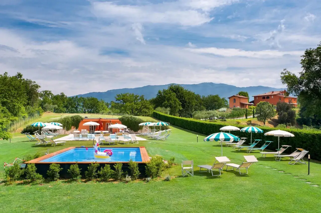 Agriturismi per bambini con piscina, lettini e ombrelloni su un prato verde, sullo sfondo colline e case in stile toscano.
