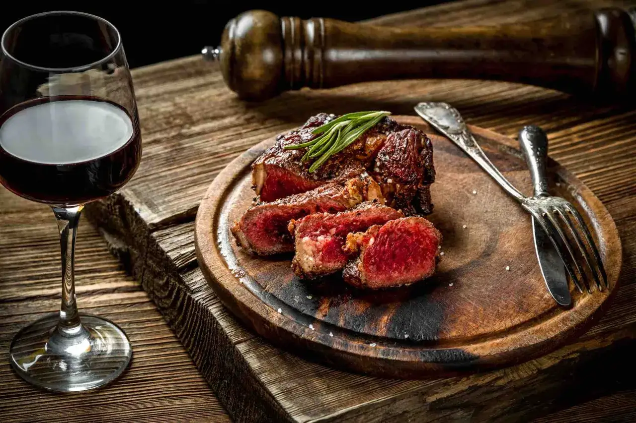 Succulenta bistecca fiorentina, tagliata e servita su tagliere di legno, con un bicchiere di vino rosso e un macina pepe sullo sfondo.