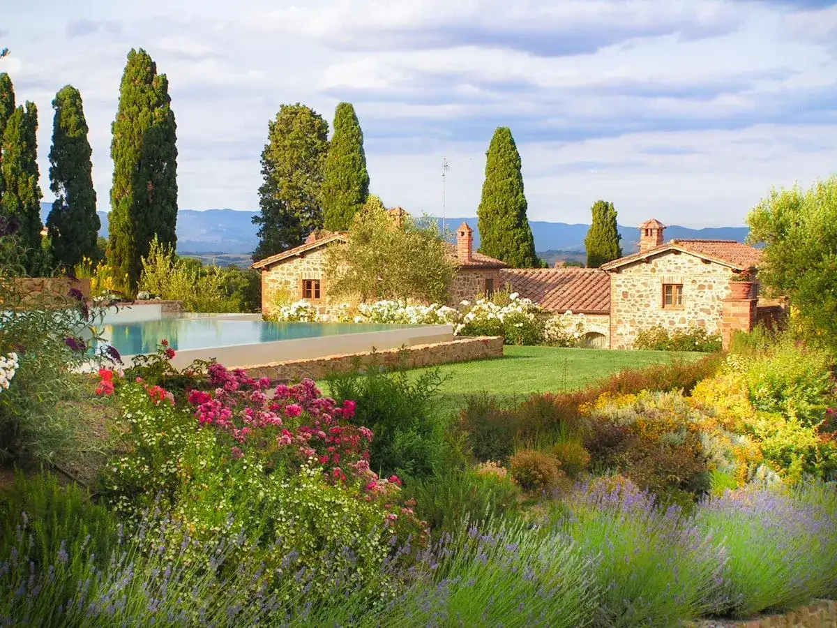 Un sogno di viaggio in Toscana: piscina, cipressi e casali in pietra tra fiori colorati e colline.