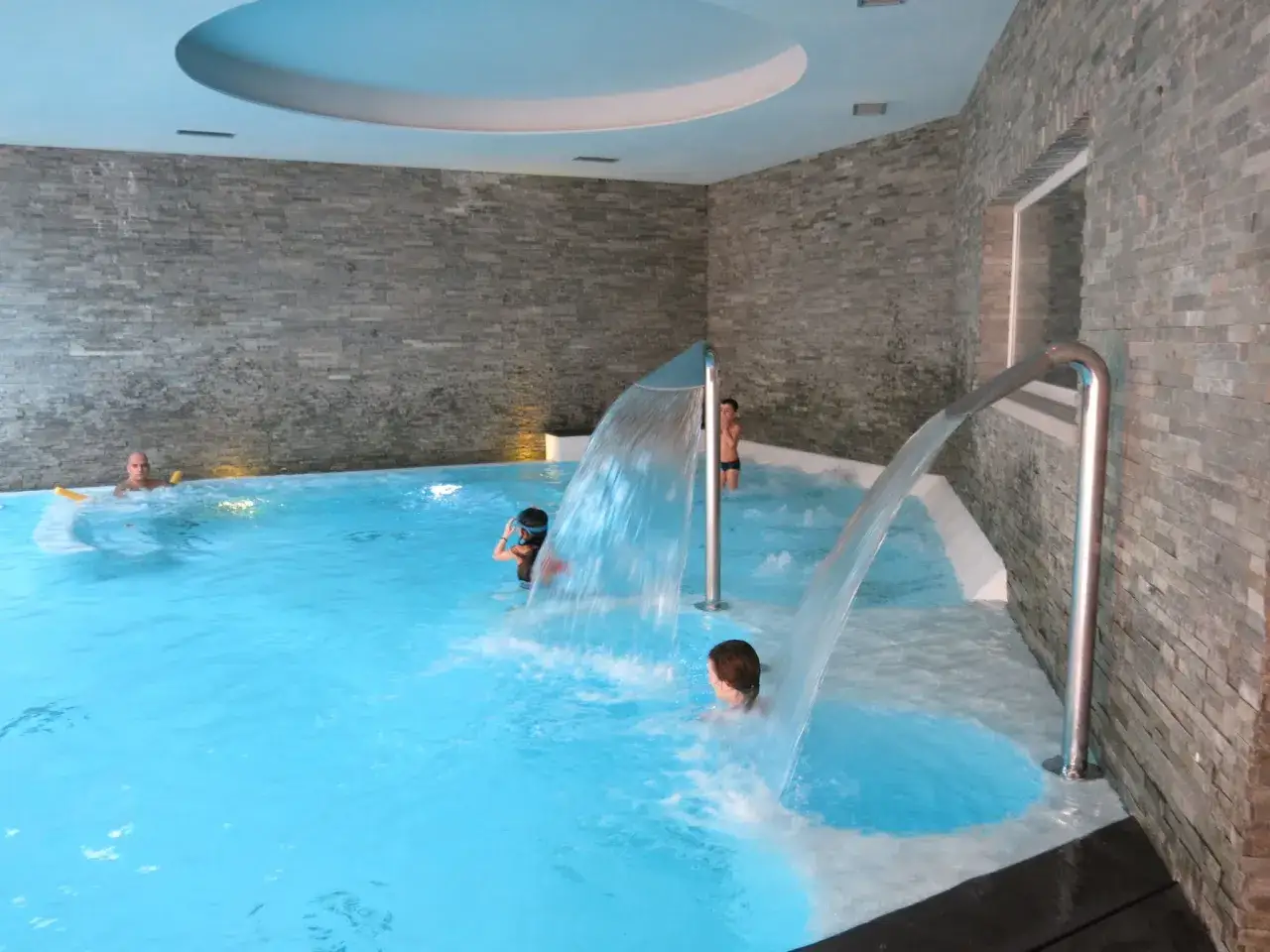 Bambini si divertono sotto cascate d'acqua in una piscina interna, perfetta spa per famiglie in Lombardia.