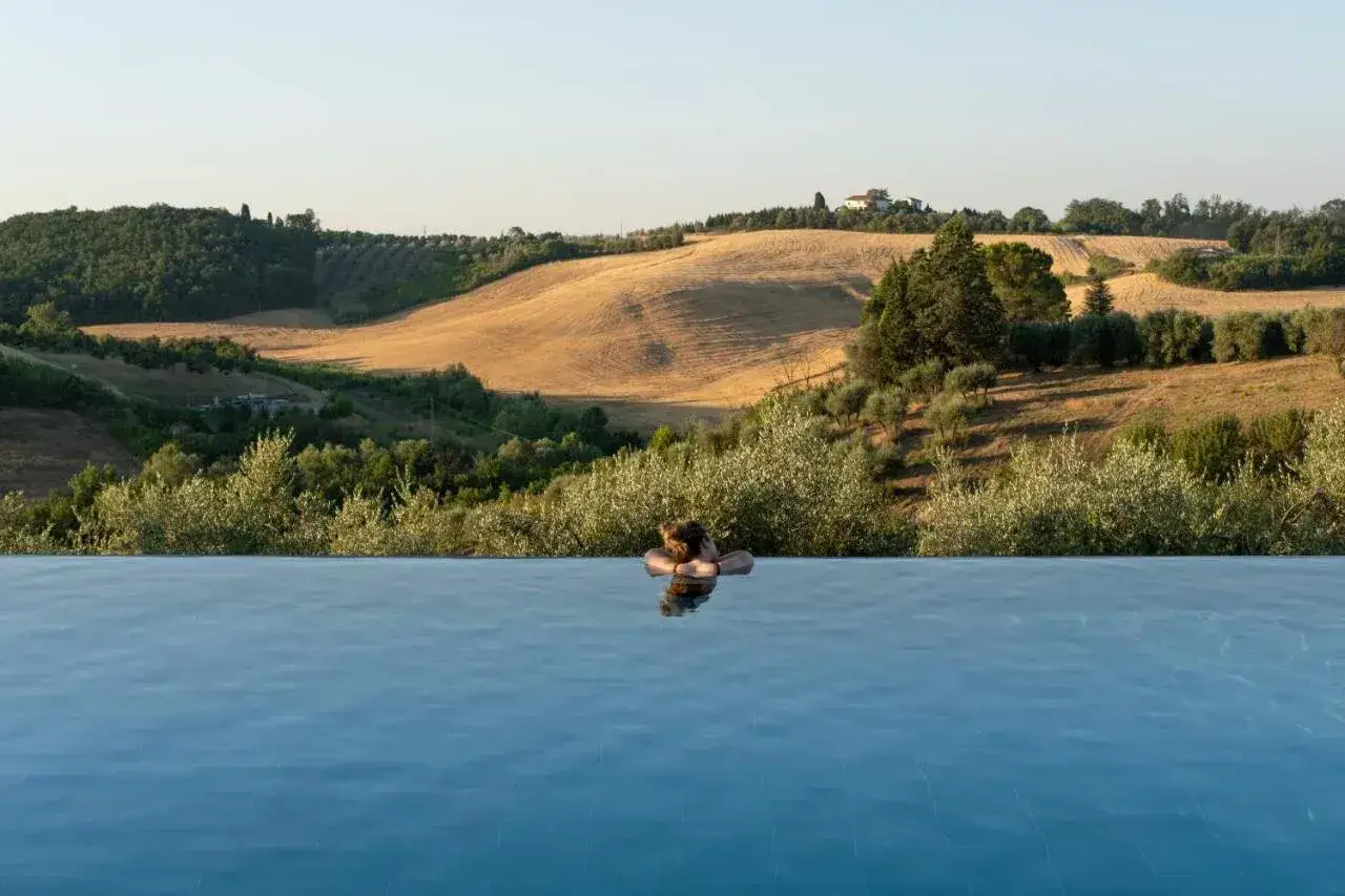 Una persona si rilassa in una piscina a sfioro con vista su dolci colline ondulate e campi dorati, un'oasi di pace in campagna.