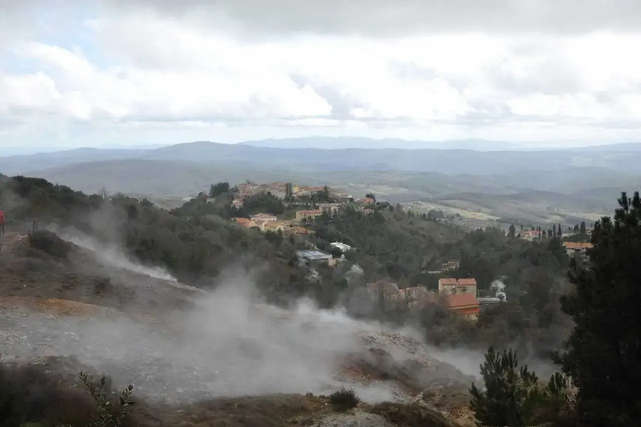 Vapore sale da un terreno brullo nel Val di Cecina, con un borgo collinare sullo sfondo.