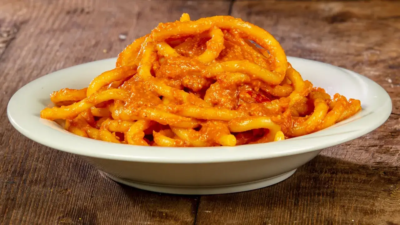 Pici al sugo d'aglione della Val di Chiana, un piatto tradizionale toscano servito in una ciotola bianca su un tavolo di legno.